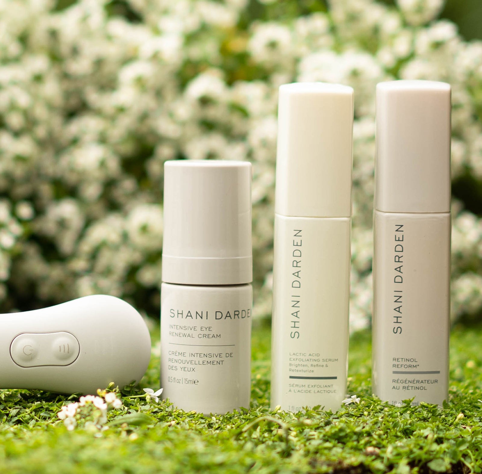 bright photo of three serums and a facial sculpting wand on a bacground of green plants with clusters of white flowers