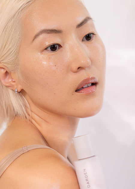 Woman with glowing, radiant skin holding Shani Darden Sake Nourishing Essence bottle against her shoulder